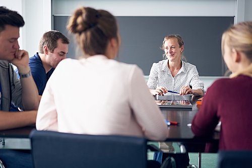 Eine Person sitzt lächelnd vor einer Tafel. Im Vordergrund sind zwei sitzende Personen von hinten zu erkennen, links sind zwei Personen von der Seite zu sehen. 